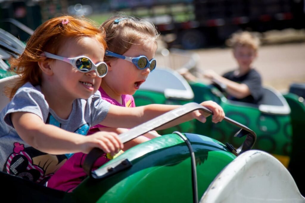 Children riding pedal go-karts on a sunny day wearing safety glasses. Bright colors and playful expressions capture the fun.