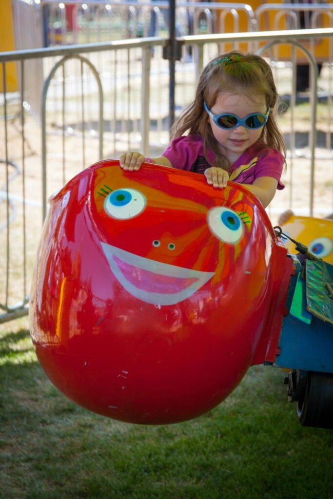 A young girl wearing sunglasses enjoys a colorful ball-shaped ride at a fair.
