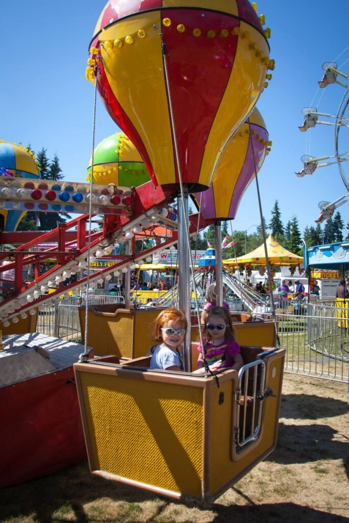 A bright red and yellow balloon ride at a fair, with a ferris wheel in the background under a clear blue sky.
