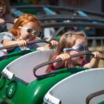 Children wearing cool sunglasses enjoy a green roller coaster ride on a sunny day.