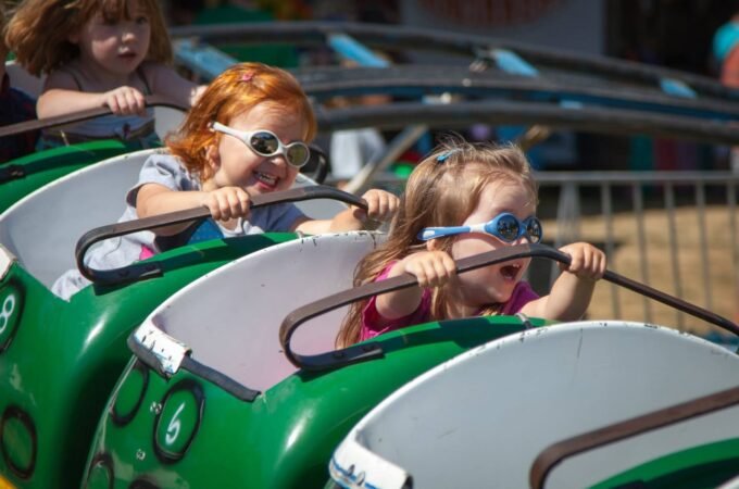 Children wearing cool sunglasses enjoy a green roller coaster ride on a sunny day.