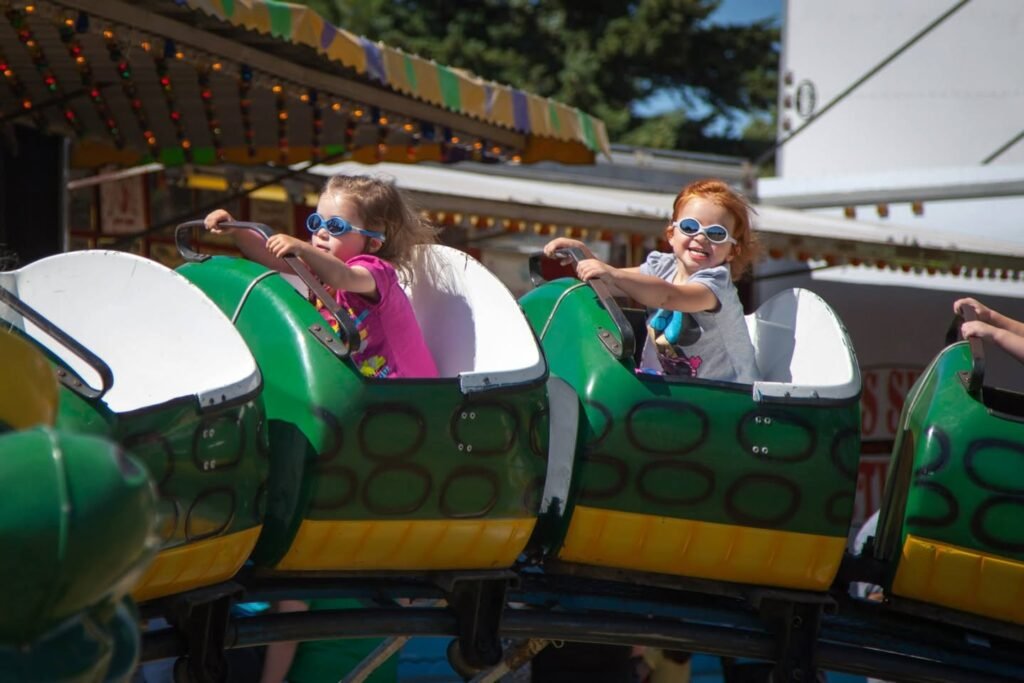 Two children enjoying a ride on a green and yellow amusement park car. They are wearing sunglasses and holding onto safety bars under a striped canopy.