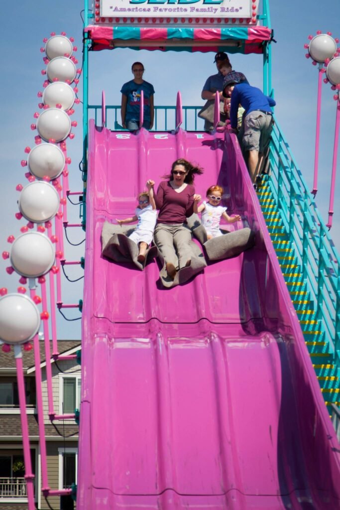 A person holds a child on a giant pink water slide at a waterfront tiki slide, with spectators watching from the top.