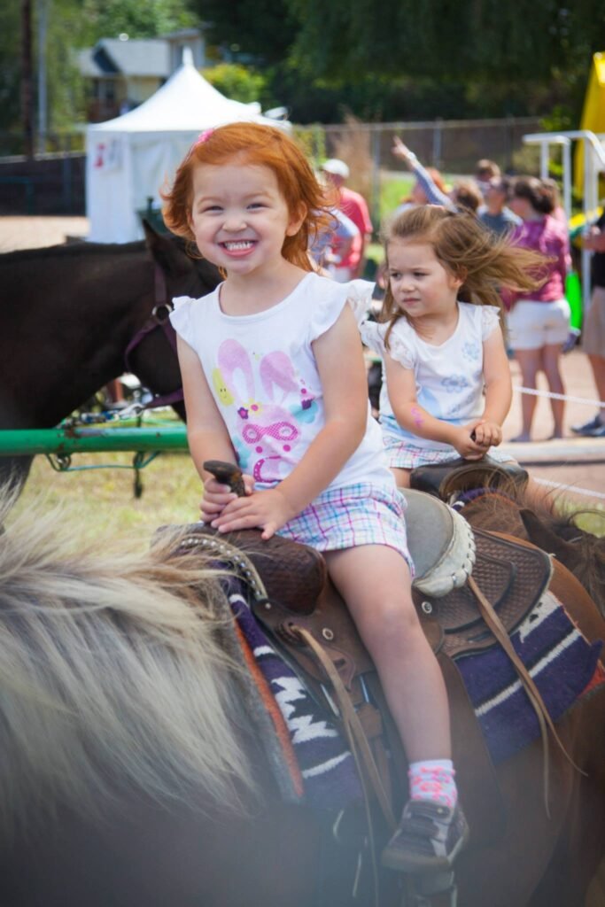 A smiling child enjoys a pony ride at a fair, with other riders and a white tent visible in the background.