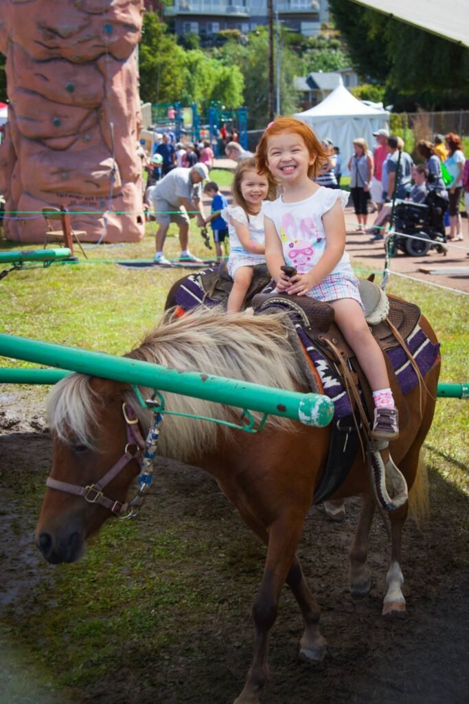 Two children enjoy a pony ride at an outdoor festival. In the background, visitors browse the community event near a rock climbing wall.