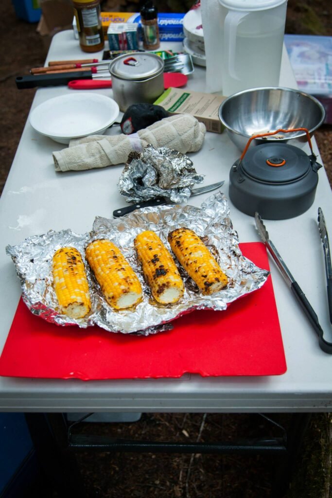 Grilled corn ears on aluminum foil with camping cookware and kettle on an outdoor table.