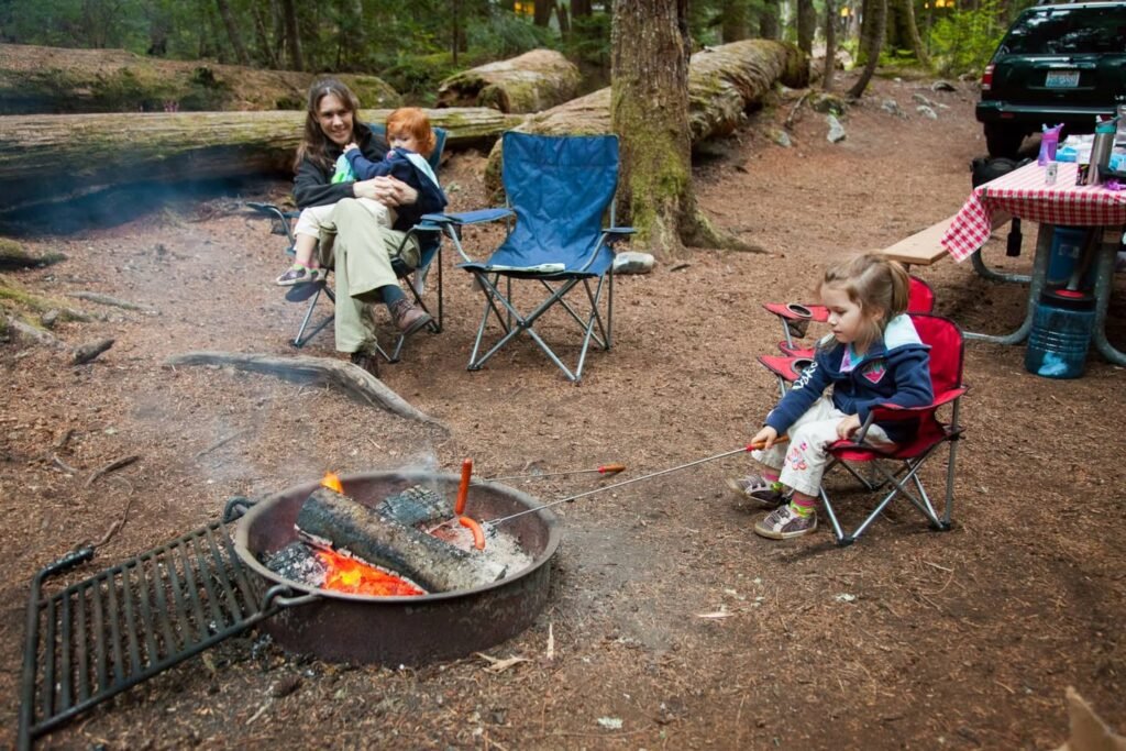 A family enjoying a warm campfire on a forested campsite with folding chairs and a red checkered picnic table.