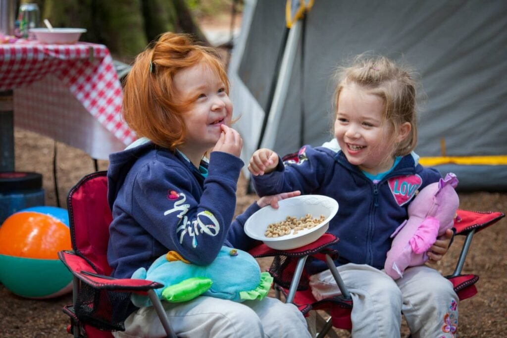 Children sitting in small camping chairs outdoors, eating from a bowl with a tent and campsite in the background.