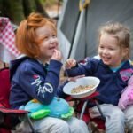 Children sitting in small camping chairs outdoors, eating from a bowl with a tent and campsite in the background.