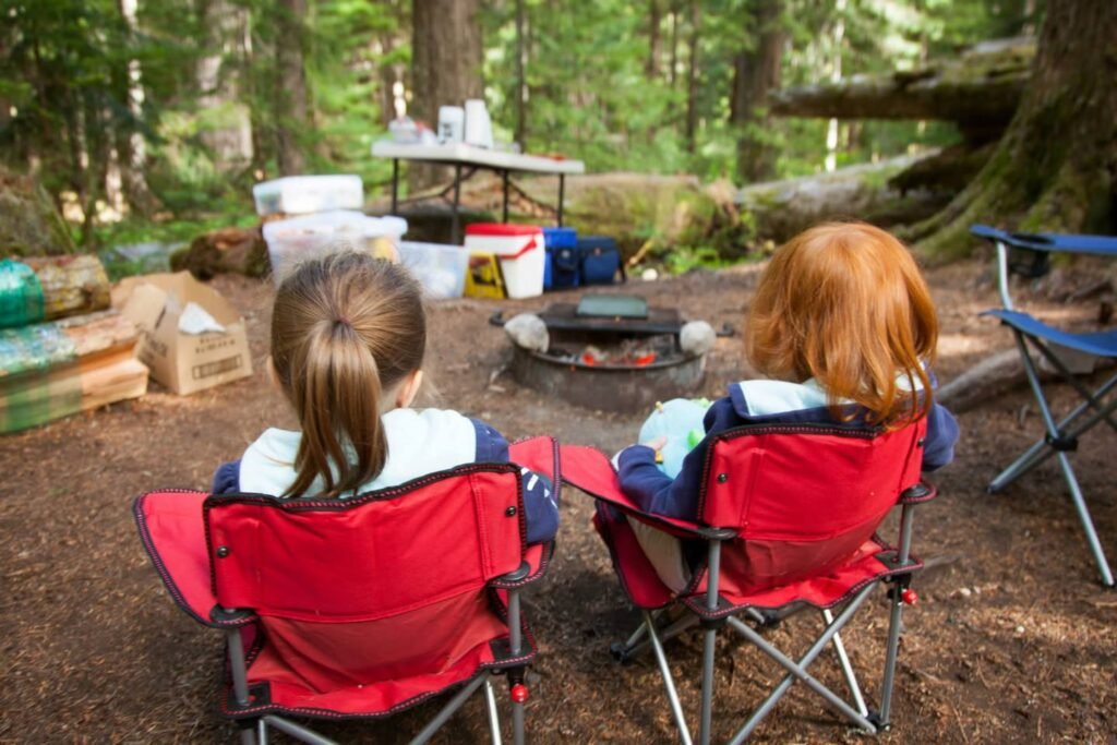 Children in red folding chairs at a wooden campground with a fire pit and coolers. The forest setting features tall evergreens and natural light.