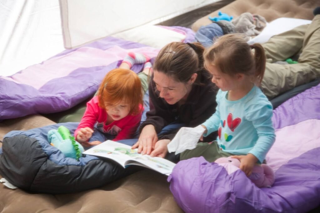 A woman and two children read a book together on sleeping bags inside a tent.