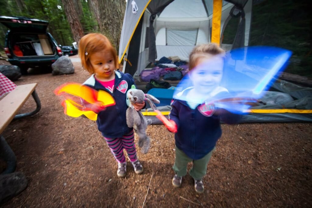 Two children holding colorful kites in a wooded campground with a family tent and vehicle in the background.