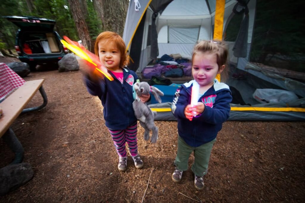 Two children holding sticks and a stuffed animal at a campsite with a tent.
