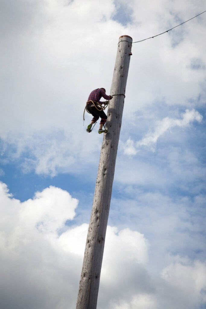 A person climbing a tall wooden pole against a partly cloudy sky. The climber is midway up the pole, with a rope visible.