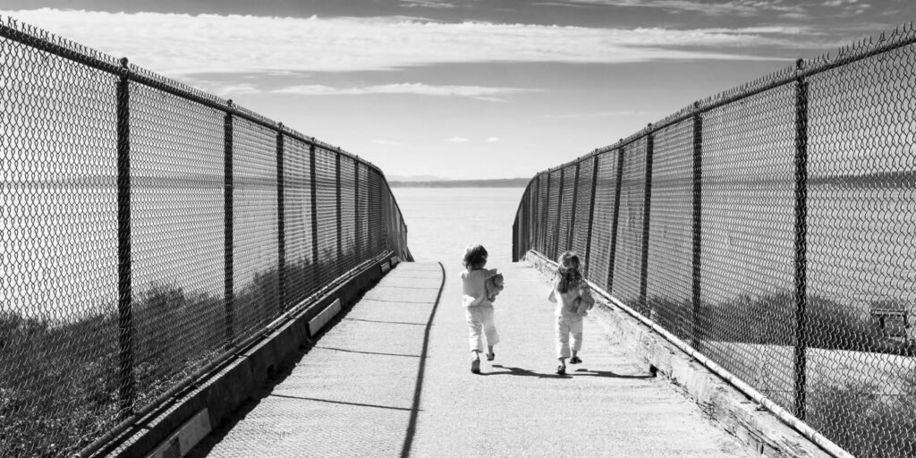 Two people walking along a curved boardwalk flanked by tall fences, with a view of the sea in the distance.
