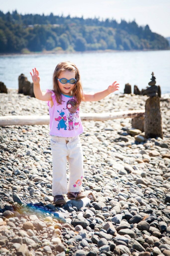 A person in a pink shirt and sunglasses plays on a rocky shoreline with old wooden pilings.