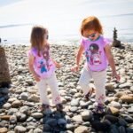 Two individuals in matching pink shirts walking on a rocky shoreline near stone cairns.