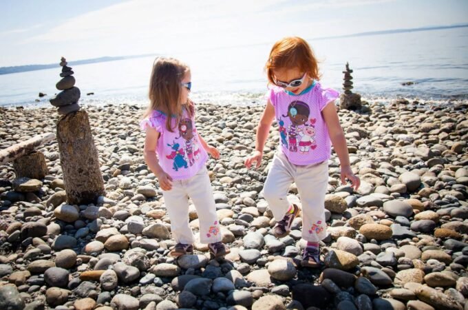 Two individuals in matching pink shirts walking on a rocky shoreline near stone cairns.