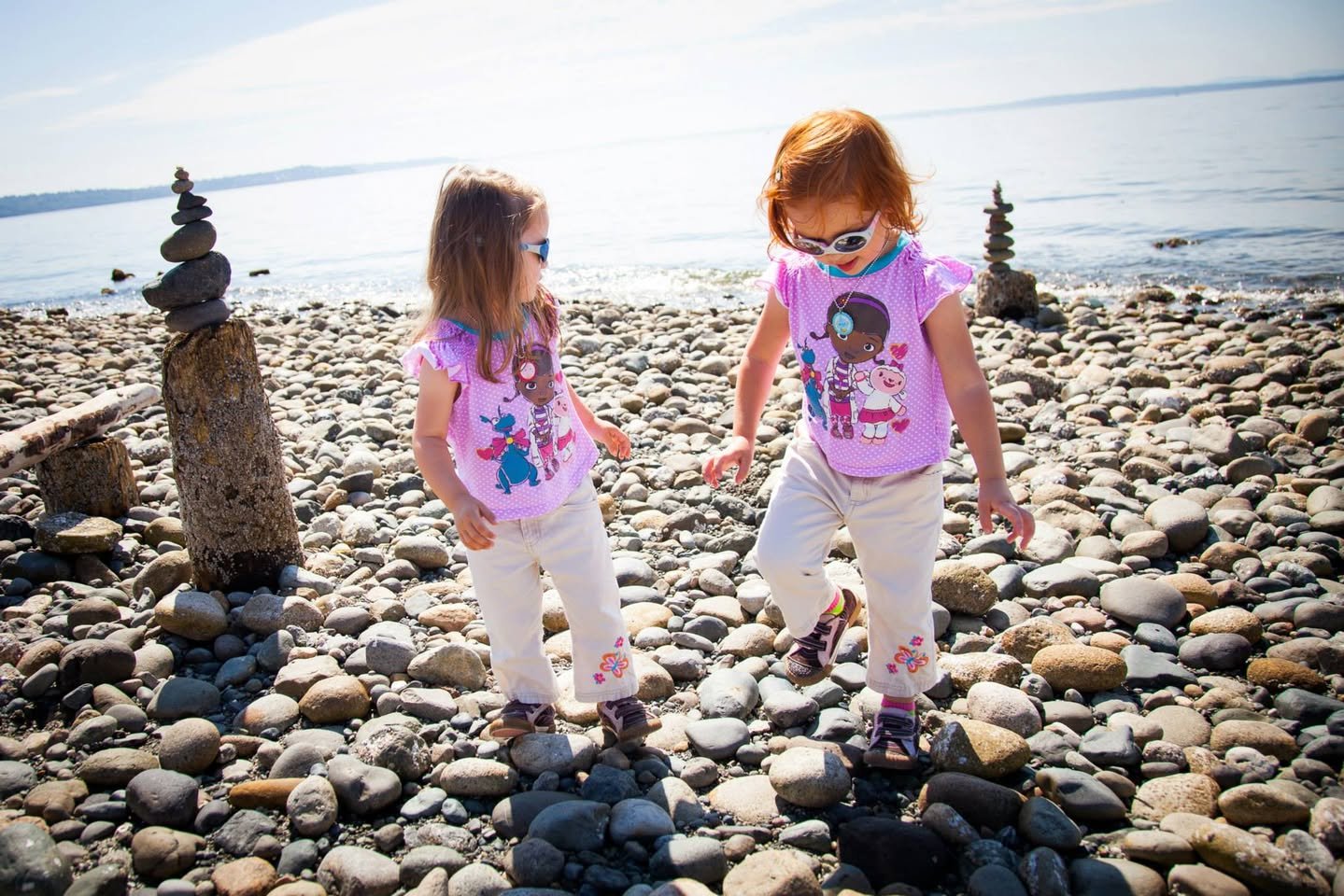 Two individuals in matching pink shirts walking on a rocky shoreline near stone cairns.