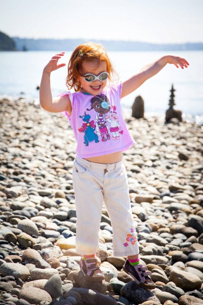 A child in sunglasses plays on a pebble beach by the water.