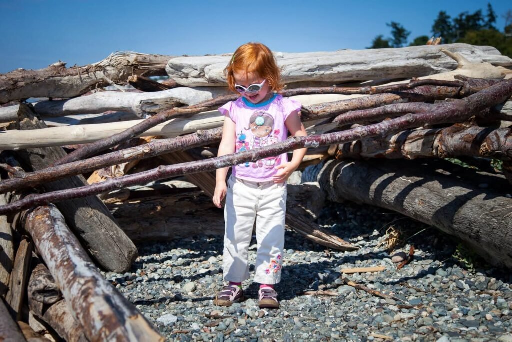A child explores a pile of driftwood and logs on a rocky shore. The scene captures a beach adventure with weathered wood under a blue sky.