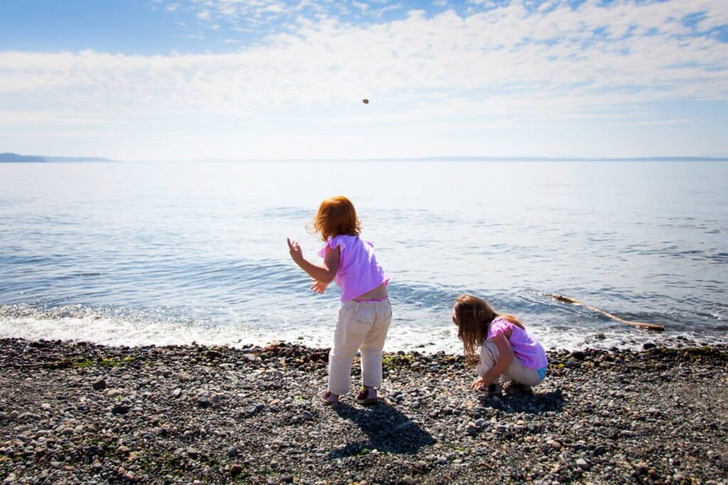 Two people enjoying a sunny day on a rocky beach in the Pacific Northwest, with one child waving a small object and another crouching near the water's edge under a bright blue sky.