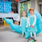 Children in matching outfits posing with a large blue inflatable whale at a bright indoor celebration.