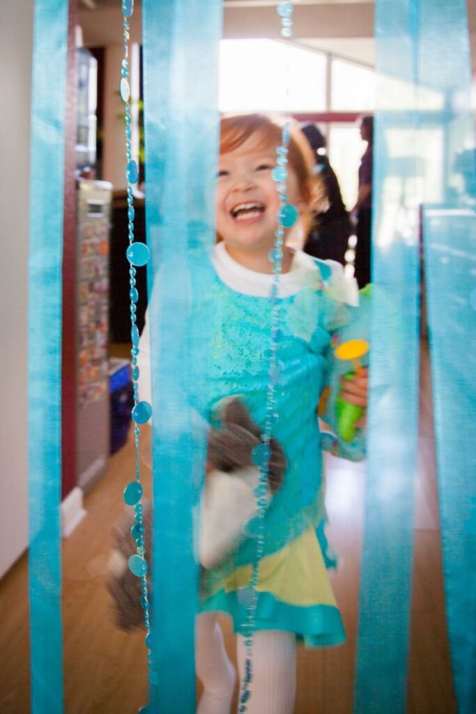 The image shows an uncropped individual wearing a bright turquoise dress with a yellow skirt, standing among hanging strings of teal beads near wooden walls and windows.