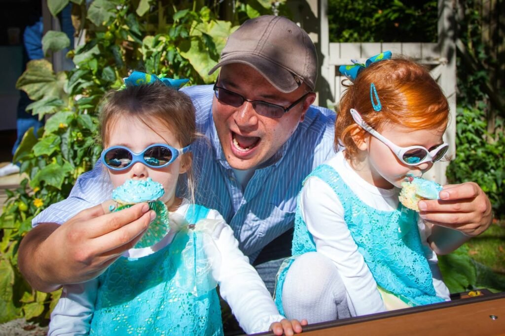 Three individuals wearing blue attire and sunglasses enjoy cupcakes outdoors. Green foliage suggests a garden setting.