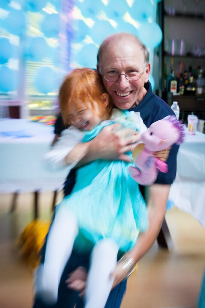 A smiling adult holds a young female child in a blue tutu dance during a celebration. The child clutches a pink stuffed unicorn toy while blue balloons decorate the background.