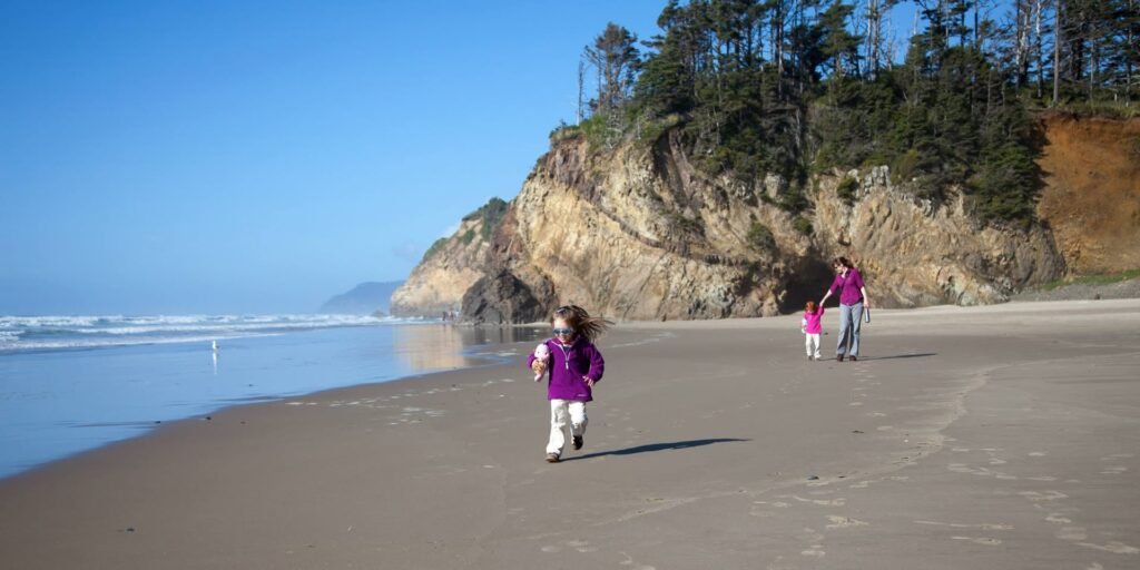 A family walks along a wide sandy beach with waves and rocky cliffs in the background under a clear blue sky.