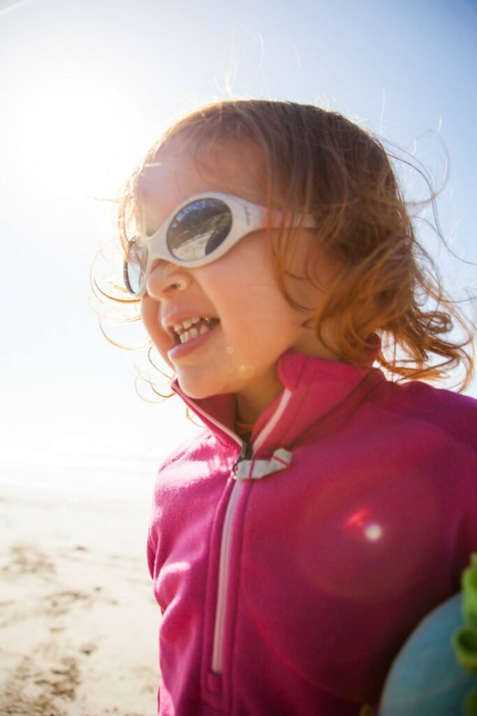 A young person in a pink zipper jacket and white sunglasses smiles on the beach with windy hair and a bright blue sky.