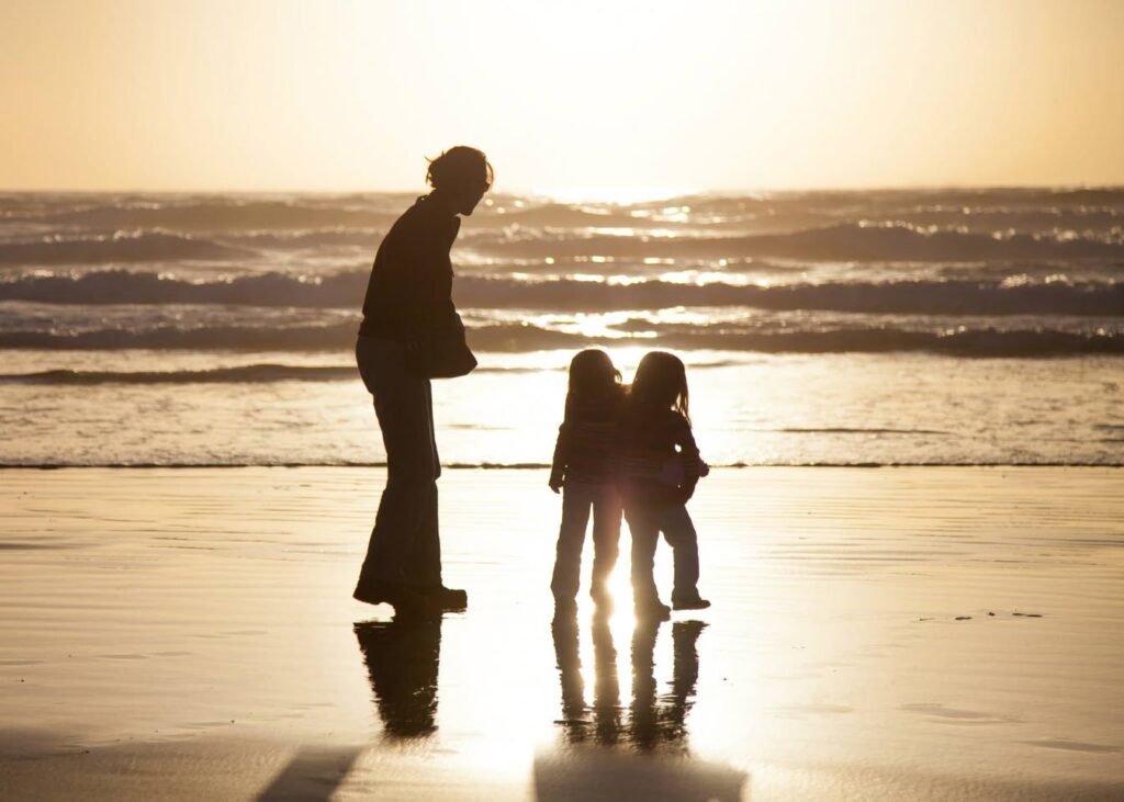 A family of three photographed as silhouettes on a wet beach at sunset. The scene captures gentle ocean waves and the golden light reflecting on the wet sand.