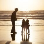 A family of three photographed as silhouettes on a wet beach at sunset. The scene captures gentle ocean waves and the golden light reflecting on the wet sand.