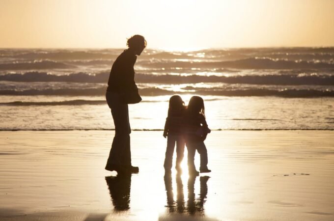 A family of three photographed as silhouettes on a wet beach at sunset. The scene captures gentle ocean waves and the golden light reflecting on the wet sand.