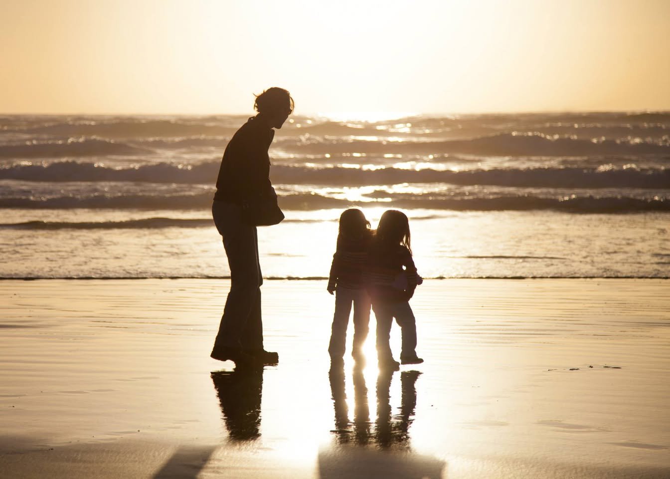 A family of three photographed as silhouettes on a wet beach at sunset. The scene captures gentle ocean waves and the golden light reflecting on the wet sand.