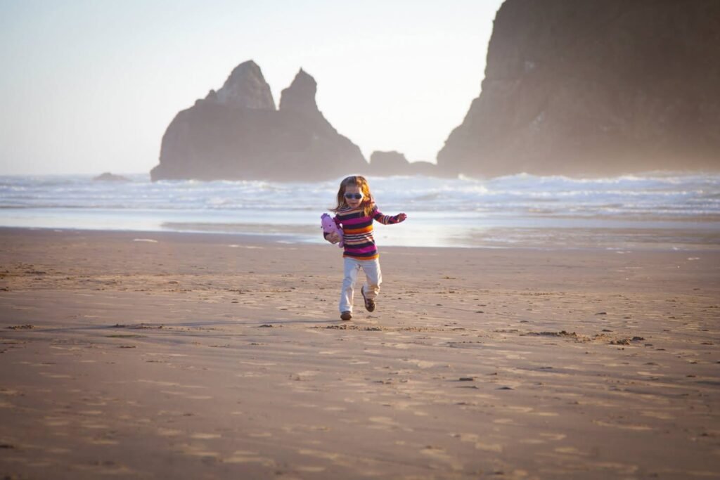 A child runs joyfully on a sandy beach with sea stacks visible in the background. The scene is bathed in soft, hazy light, creating a serene and peaceful atmosphere.