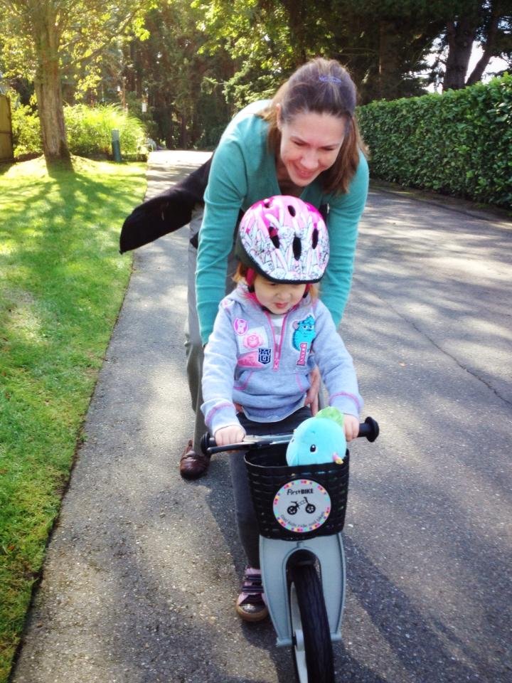 A woman in a green shirt kneels behind a toddler in a pink helmet, guiding a bike on a shaded walkway.