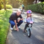 A father crouching beside his daughter as she rides her bike on a sunny path.
