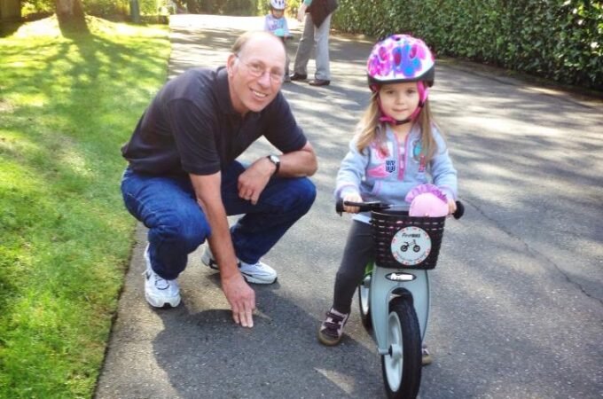 A father crouching beside his daughter as she rides her bike on a sunny path.
