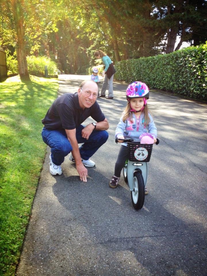 A father crouching beside his daughter as she rides her bike on a sunny path.