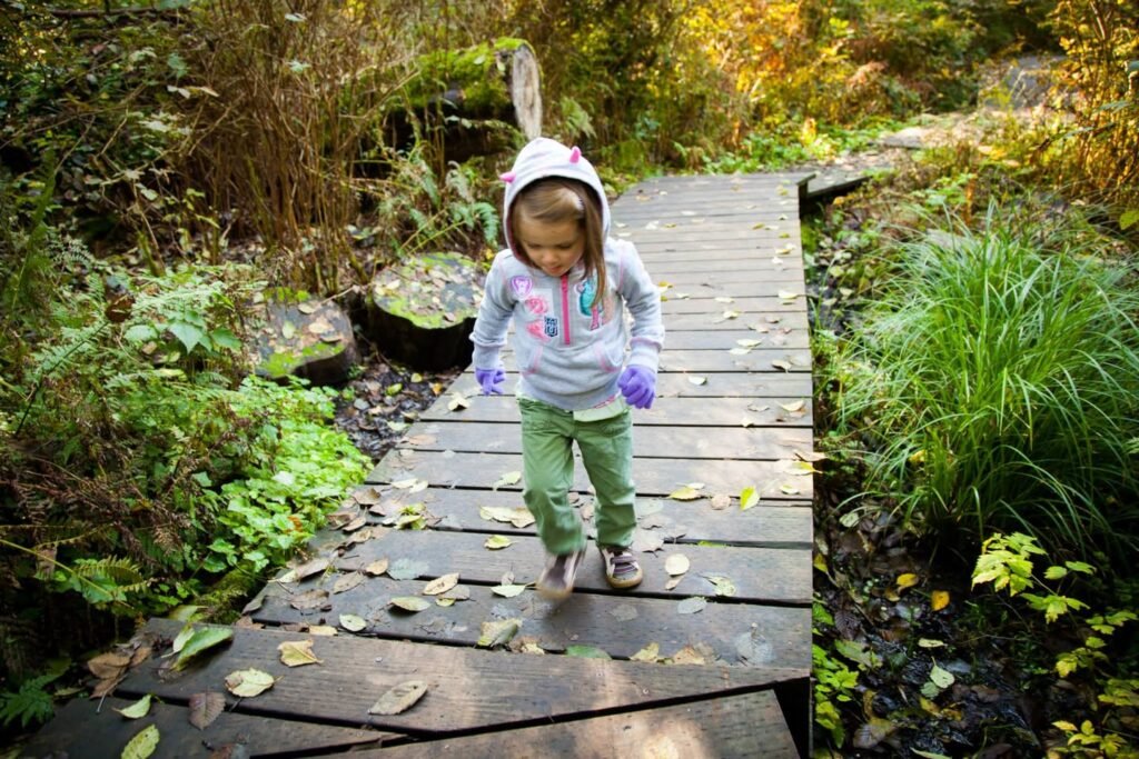 A child walks along a wooden boardwalk through a lush garden with green foliage and autumn leaves. The setting features natural pathways and outdoor scenery.