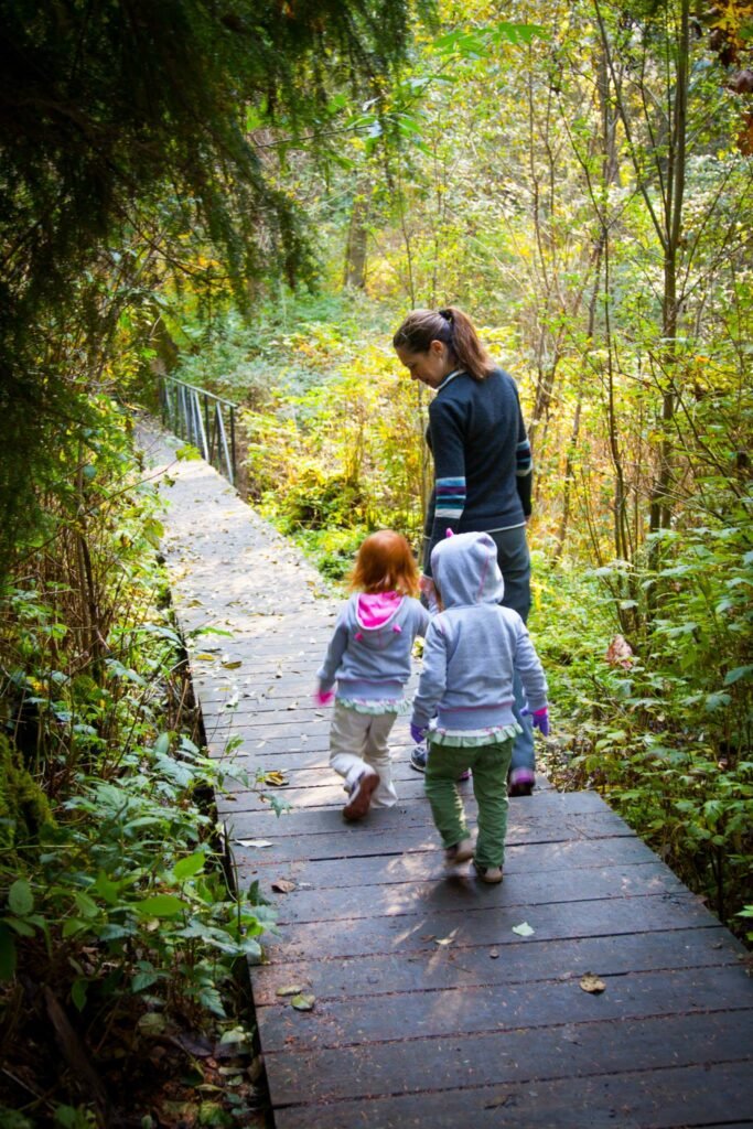 An adult and two children walking on a wooden boardwalk through a lush green forest. The scene is packed with vibrant green foliage and a clear hiking path.