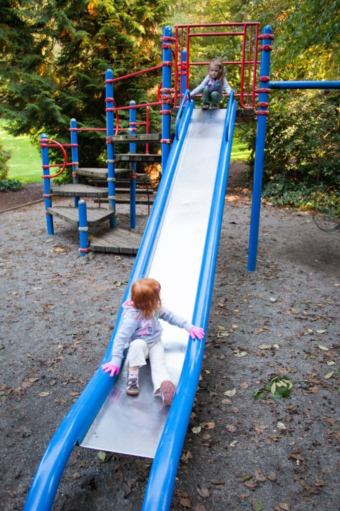 Two children slide down a long blue and silver playground slide in a green park setting.