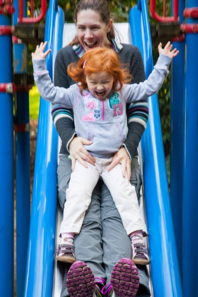 A joyful moment as a child slides down a blue playground slide while an adult holds on for fun. Bright colors and smiles capture the excitement of outdoor playtime.