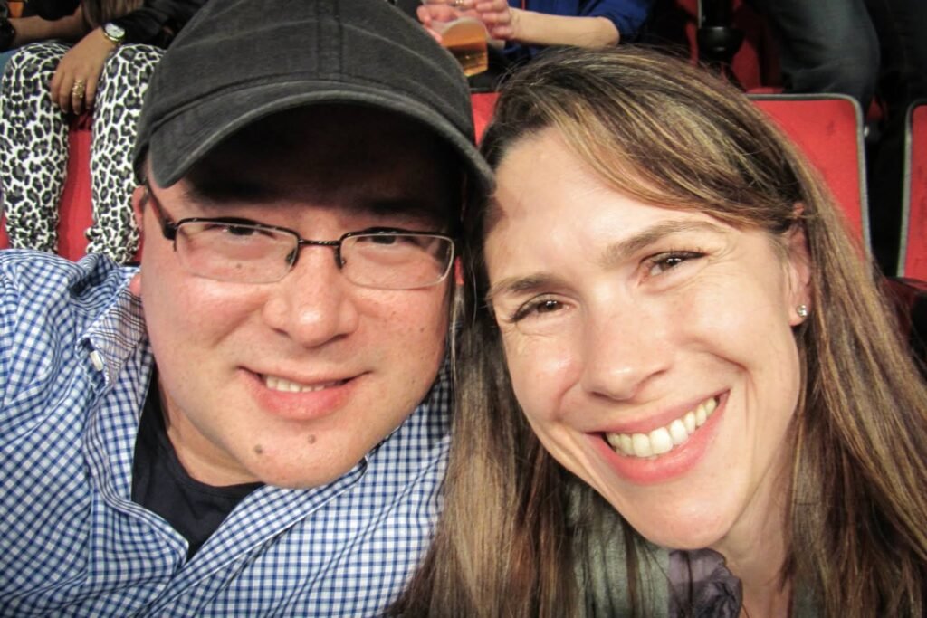 A couple smiles at the camera while seated in red theater seats during a live event.