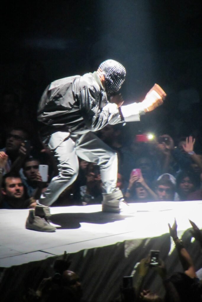 A performer at an indoor concert engages a large crowd by holding a small object while standing on an illuminated white runway stage.