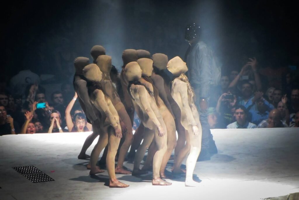A group of performers in earth-toned costumes and headscarves on a lit stage in a darkened theater.