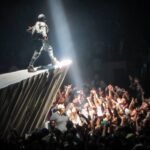 A performer on a stage wing under a spotlight above a large concert crowd with raised hands.