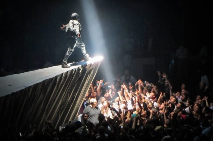 A performer on a stage wing under a spotlight above a large concert crowd with raised hands.